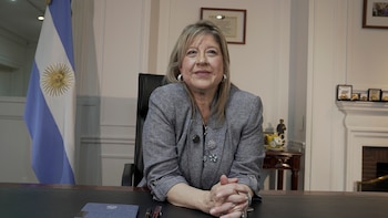 Mujer rubia de mediana edad, vestida con blazer gris y broches, sentada en un escritorio con las manos entrelazadas, con una bandera argentina detrás