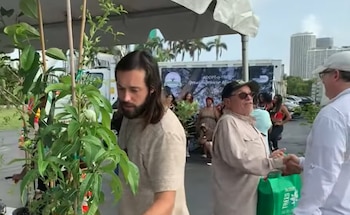Varias personas conversan bajo una carpa blanca junto a plantas en macetas y un cartel del programa Adopt-a-Tree