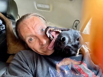 Un hombre dentro de un automóvil junto a un perro de pelaje oscuro que mantiene el hocico abierto al bostezar