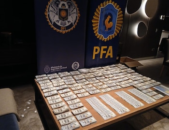 Billetes de cien dólares apilados ordenadamente sobre una mesa de madera, con dos pancartas azules de la Policía Federal Argentina (PFA) al fondo