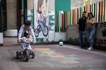 Una niña en triciclo rojo es empujada por un hombre en un patio con una rayuela pintada. Una pareja observa de fondo junto a una valla de postes coloridos