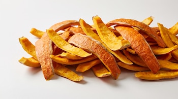Tiras y fragmentos irregulares de cáscaras de calabaza fresca de color naranja intenso, amontonadas sobre una superficie blanca.