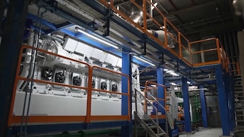 Gran máquina industrial blanca con detalles naranjas y azules, tubos y ventiladores visibles, dentro de una fábrica con pasarelas metálicas y escaleras