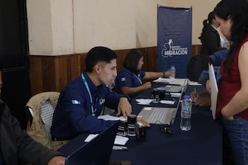 Funcionarios en uniforme azul atienden a personas frente a computadoras y sellos junto a un cartel del Instituto Guatemalteco de Migración
