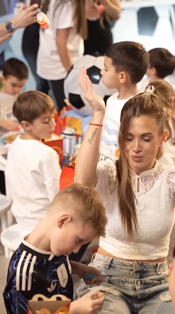 Mujer con blusa blanca y jeans rasgados, tatuaje en el brazo. Varios niños, uno con camiseta de fútbol, mesas con cajas de comida y un balón.