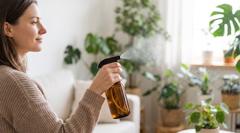 Mujer de perfil con suéter de punto marrón rociando un líquido de una botella de vidrio ámbar. Se ve el rocío y varias plantas de interior al fondo.