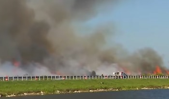 Vista panorámica de un gran incendio forestal con humo denso y llamas, una carretera con barrera, césped y agua, y un vehículo de emergencia a lo lejos