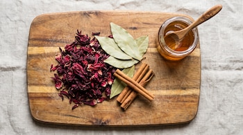 Flores de jamaica secas, hojas de laurel, ramitas de canela, frasco de miel y cuchara de madera sobre tabla de cortar y mantel de lino.