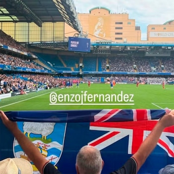 Persona de espaldas sostiene bandera de las Islas Malvinas y Reino Unido en estadio. Jugadores en cancha, público en gradas y pantalla electrónica
