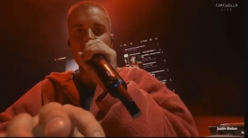 Primer plano de Justin Bieber con cabello rapado, micrófono en mano y sudadera roja, actuando en un escenario oscuro con luces rojas y una pantalla al fondo