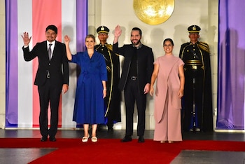 Cuatro personas con vestimenta formal saludan con la mano alzada frente a dos guardias de honor y un emblema dorado en la pared