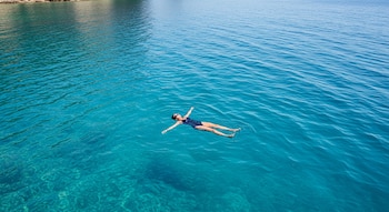 Vista aérea de una persona flotando boca arriba en un traje de baño oscuro en aguas marinas cristalinas de color azul turquesa, cerca de una costa rocosa.