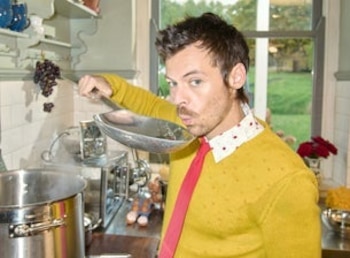 Hombre en una cocina, viste suéter amarillo, camisa blanca con puntos rojos y corbata roja, sostiene una cuchara grande sobre una olla. Hay uvas colgando y una ventana