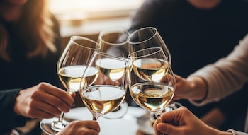 Cuatro manos sujetan copas de vino blanco, chocando en un brindis. Personas con ropa oscura y clara se ven desenfocadas en el fondo.
