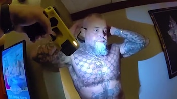 Un hombre barbudo y tatuado, sin camisa, levanta las manos mientras es iluminado por un dispositivo amarillo brillante. Se ve una pantalla a la izquierda