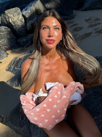 Una mujer de cabello largo sostiene a un bebé envuelto en una manta rosa con estampado sobre un fondo de rocas y arena
