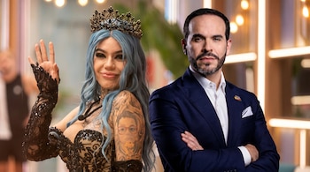 Una mujer con cabello azul, corona negra y tatuajes posa con un hombre barbudo en traje azul marino y camisa blanca con los brazos cruzados.