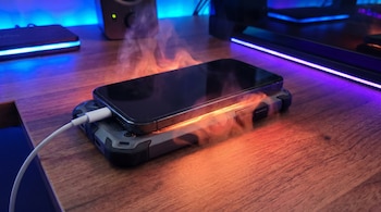 Smartphone moderno cargándose en una mesa de madera con una funda gruesa. Efecto visual de calor y vapor se eleva alrededor del dispositivo, con luces tecnológicas.