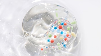 Gel transparente y viscoso con burbujas de aire y múltiples microcápsulas esféricas de colores rojo, azul, verde y amarillo sobre un fondo blanco.