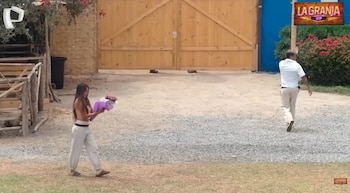 Una mujer de cabello largo oscuro, vestida de claro, camina en un sendero de grava sosteniendo un ramo de flores envuelto en morado. Un hombre y una puerta de madera al fondo
