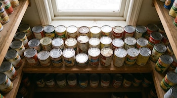 Numerosas latas de alimentos de diferentes marcas y tamaños se apilan en estantes de madera bajo una ventana; algunas tapas muestran óxido.