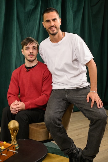 Dos hombres sonriendo: uno sentado con suéter rojo, otro de pie con camiseta blanca. Fondo de cortina verde. Trofeo de la Copa del Mundo y futbolín