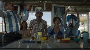 Cuatro adultos, tres hombres y una mujer, con expresiones de asombro y euforia, sentados en la mesa de un restaurante adornado con colores argentinos. Hay vasos y platos