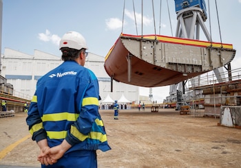 Hombre con casco de seguridad y uniforme azul en primer plano observa un bloque de casco de barco levantado por una grúa en un astillero