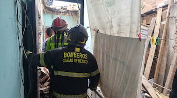 Vista trasera de dos bomberos de la Ciudad de México con cascos y uniformes. Pared azul, cables eléctricos, tendedero con sábanas y ladrillos expuestos.