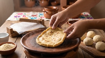 Dos manos forman el borde de un disco de maíz sobre un comal de barro, junto a bolas de masa y una prensa de madera sobre una mesa.