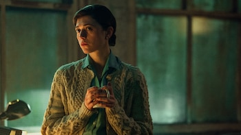 Una mujer de cabello oscuro con camisa verde y cárdigan beige sostiene una taza blanca. Su rostro está iluminado con luz cálida, mirando a la izquierda