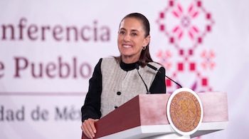 Mujer con cabello recogido, chaqueta texturizada, sonríe y habla en un podio con un micrófono y el escudo de México, frente a un fondo blanco con texto borroso.