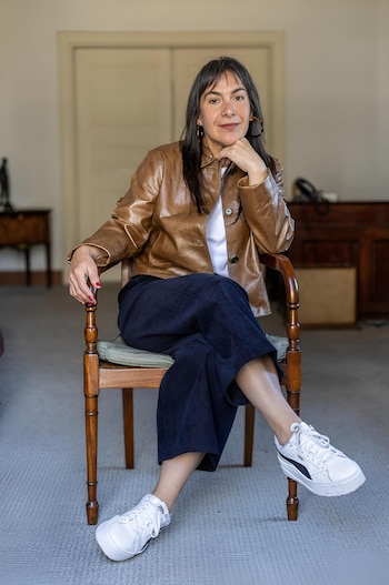 Retrato de cuerpo entero de Tania Candiani, sentada en una silla de madera, con chaqueta de cuero marrón, pantalón oscuro y zapatillas blancas