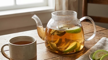 Tetera de cristal con infusión de hojas y rodajas de guayaba humeante sobre mesa de madera, con una taza de té y más rodajas de fruta al lado, frente a una ventana.