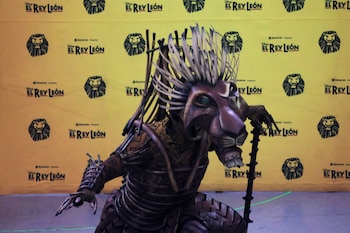 Figura con máscara de león y bastón frente a un fondo amarillo con logotipos repetidos del musical El Rey León