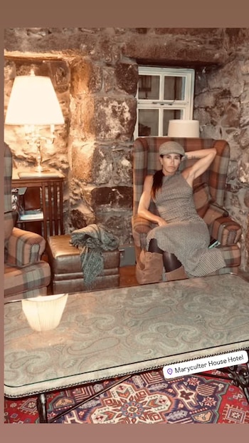 Carolina Baldini con vestimenta a cuadros y boina sentada en un sillón junto a una pared de piedra
