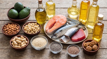 Mesa de madera con aguacates, almendras, anacardos, maní, semillas de sésamo, chía, lino, nueces, salmón, atún, caballa, sardinas y botellas de aceite.