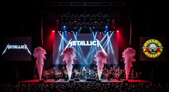 Escenario de concierto con orquesta sinfónica, humo ascendente, luces dramáticas rojas y azules, y los logos de Metallica y Guns N' Roses proyectados. Asientos de audiencia visibles.