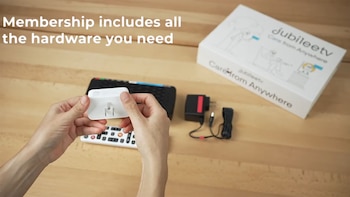 Una persona sostiene un adaptador blanco con clavijas sobre una mesa de madera. En el fondo, se observan dos controles, un cargador, cables y la caja de JubileeTV