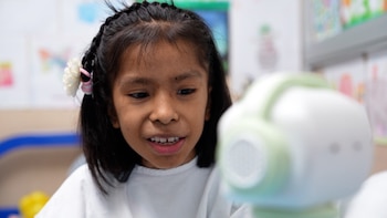 Primer plano de una niña con cabello oscuro y trenzas, interactuando con un robot blanco y verde. La niña mira hacia abajo, con la boca entreabierta