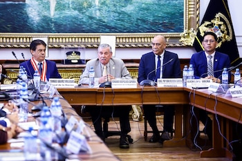 Cuatro hombres sentados en una mesa de madera en una sala formal. Hay micrófonos, botellas de agua y carteles de identificación frente a cada persona. Un cuadro grande cuelga detrás