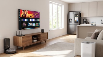 Un televisor inteligente, un enrutador, un robot aspirador, un purificador de aire y un refrigerador con pantalla en un salón con cocina.
