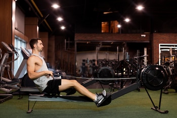 Un hombre vestido con camiseta sin mangas gris y pantalones cortos negros utiliza una máquina de remo estático en un gimnasio con equipo deportivo