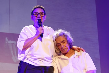 Retrato de una mujer sonriente con pelo corto oscuro y gafas redondas negras, vestida con una blusa fucsia y naranja, con las manos entrelazadas sobre una mesa de madera
