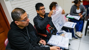 Varias personas sentadas en pupitres examinan documentos electorales dentro de bolsas transparentes en un aula de clases