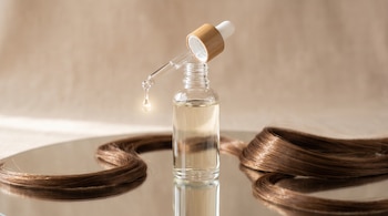 Frasco de vidrio con gotero vertiendo una gota de producto transparente sobre una superficie reflejante junto a mechones de cabello castaño.