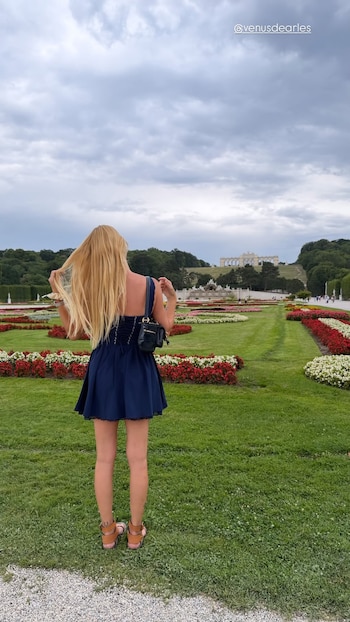 Mujer rubia de espaldas con vestido azul, sandalias y bolso negro, de pie en un jardín con flores rojas y blancas, árboles y un palacio al fondo bajo cielo nublado