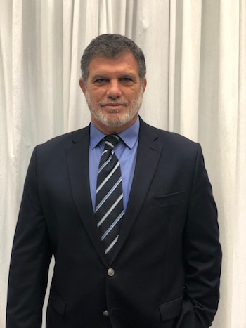 Hombre canoso con barba, traje azul oscuro, camisa de vestir y corbata a rayas delante de una cortina blanca