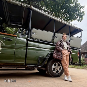 Una mujer, un vehículo de safari verde con techo de lona, grandes ruedas, un árbol, vegetación, y la palabra Addos en el suelo