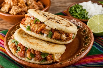 Dos gorditas de maíz rellenas de chicharrón en un plato de barro, junto a recipientes con cilantro, cebolla y medio limón.